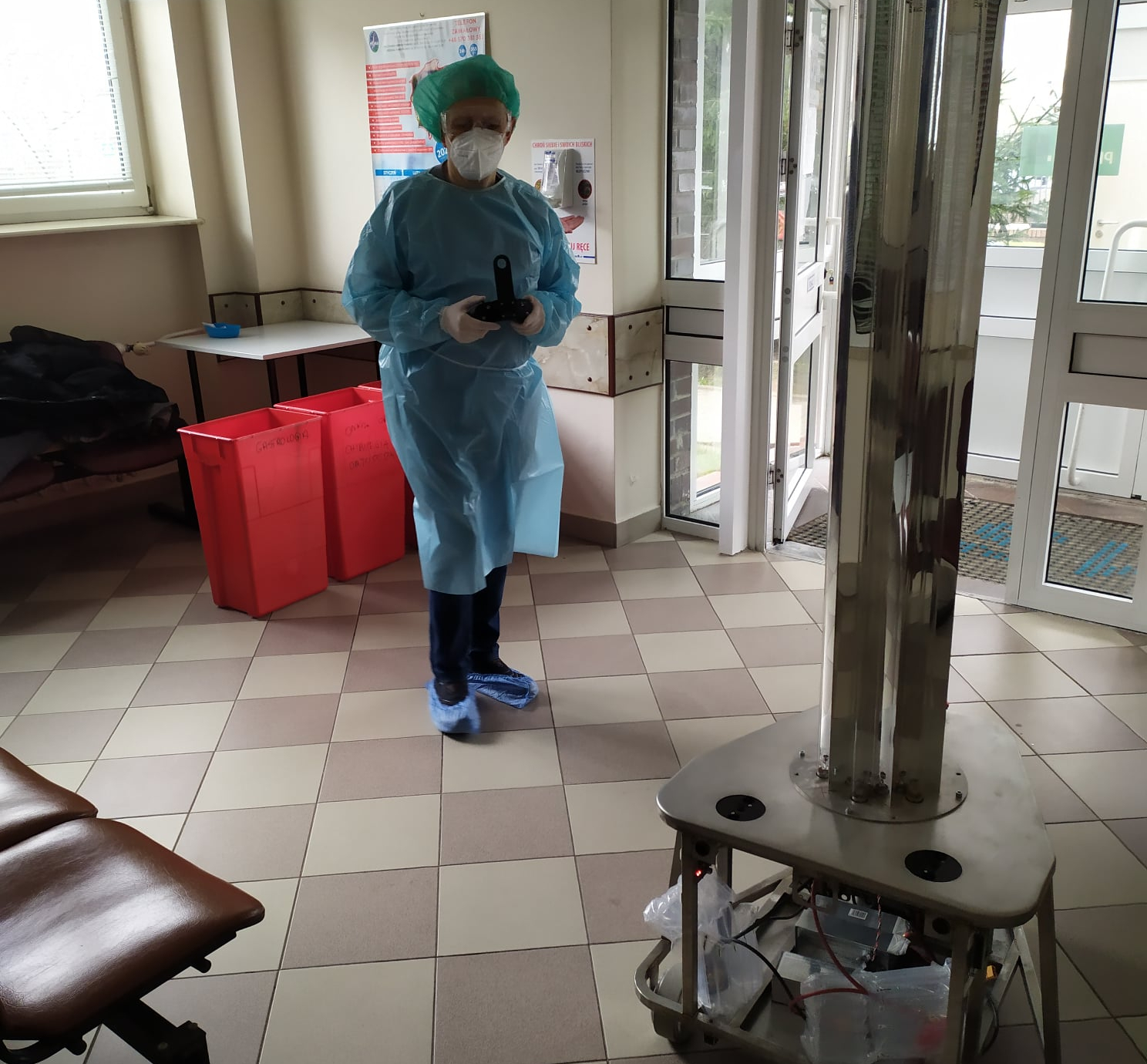 Korytarz szpitalny. Mężczyzna w fatruchu i masce, w ręku kontroler. Przed nim samobieżny robot.